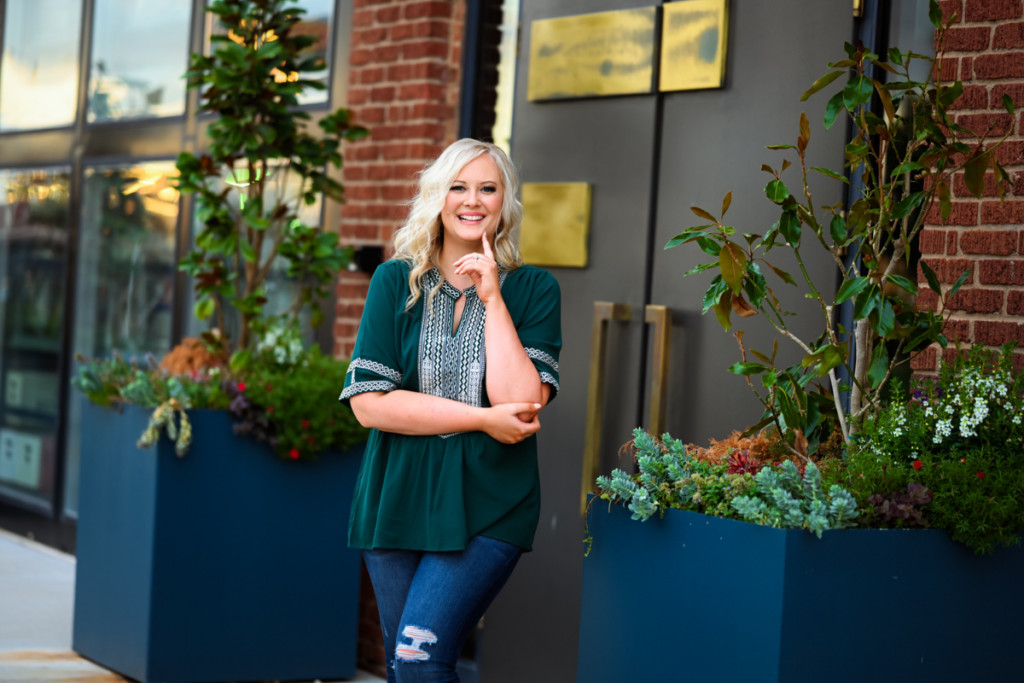 A blonde woman standing in front of a building with potted plants, using social media for customer insights.