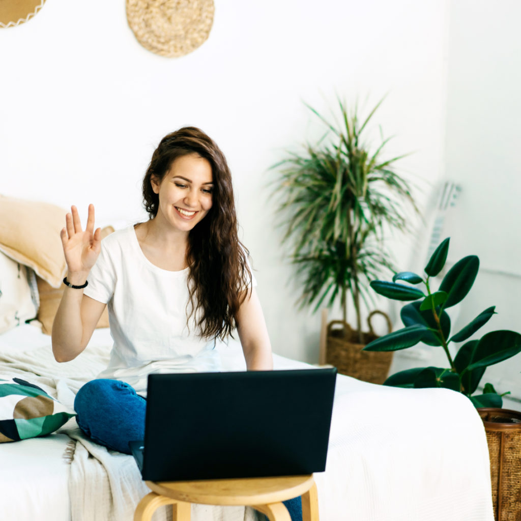 A young woman, a mastermind, sitting on a bed with a laptop and waving.