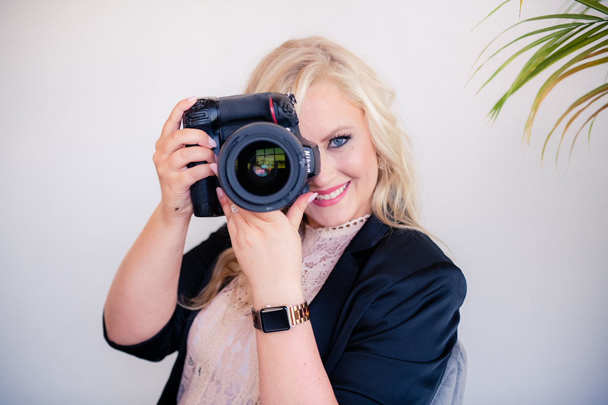 Blonde woman in black blazer and pink shirt holding a camera up to her eye