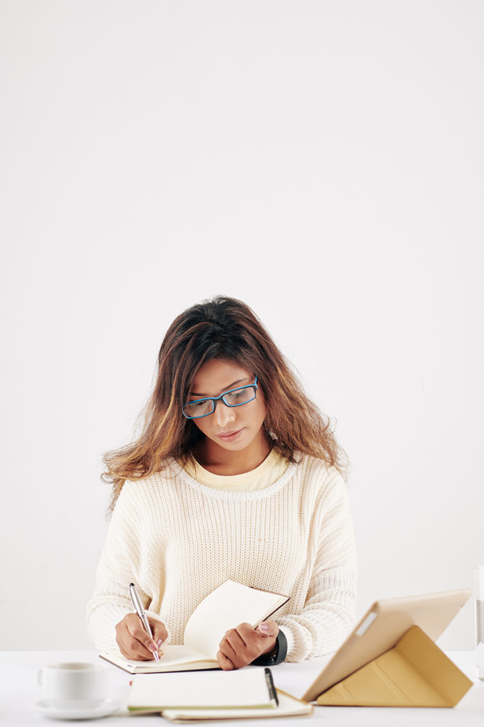 A woman wearing glasses is sitting at a desk and writing in a notebook, while utilizing business marketing tips.