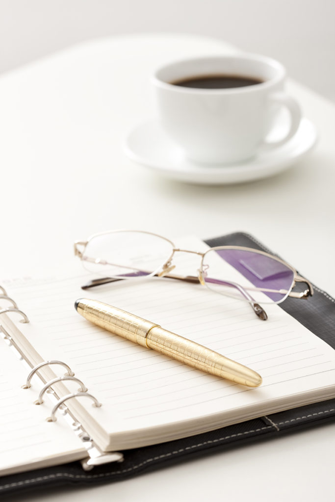 A notebook with a cup of coffee and a pen on a white table, perfect for jotting down business marketing tips.