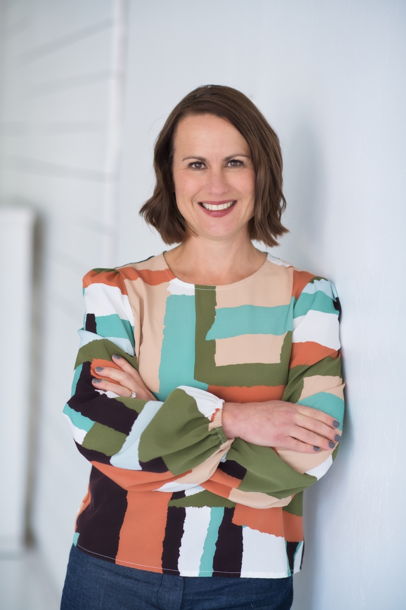 A woman in a colorful top standing against a wall.