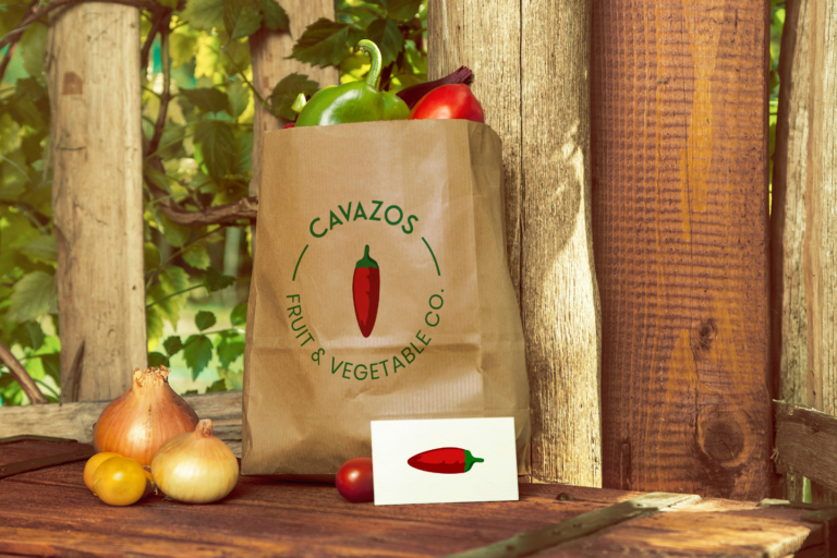 A paper bag branded with "Cavazos Fruit & Vegetable Co." is filled with fresh produce, with additional vegetables scattered on a wooden surface.