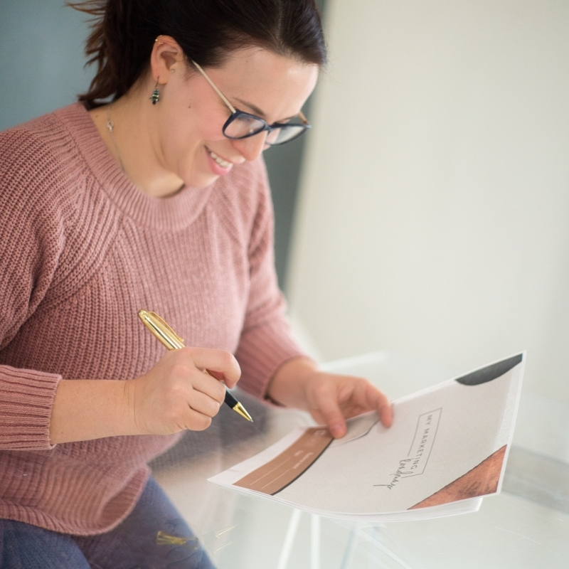 A smiling woman in glasses and a pink sweater reviews an e-learning document and writes notes at a glass table.