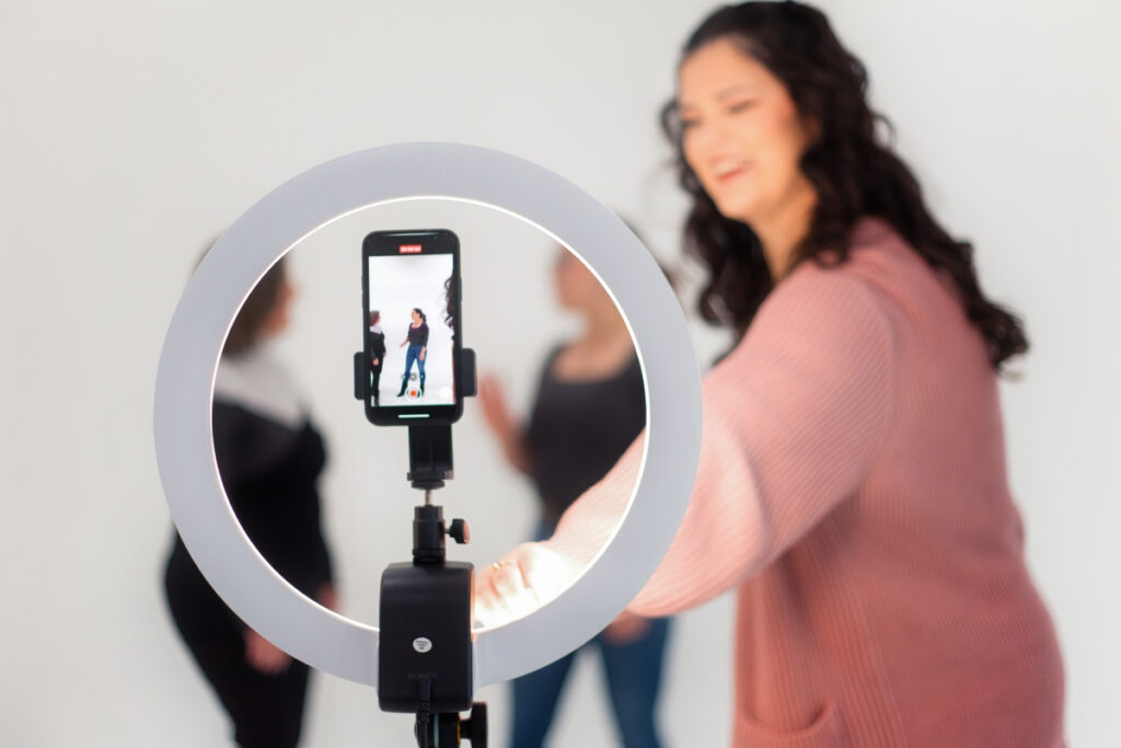 A woman adjusting a ring light while recording a social media marketing video with her smartphone. Two women are seen in the background.