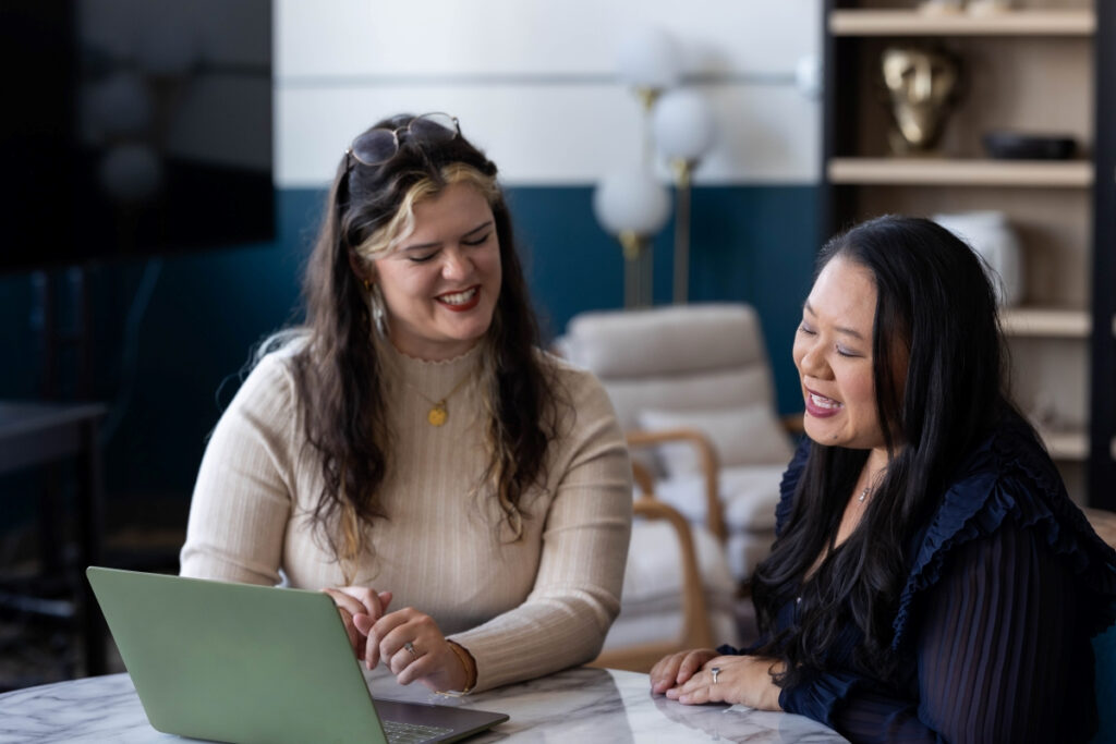 Two women smiling and working on a laptop during a meeting, discussing Black Friday sale strategies.