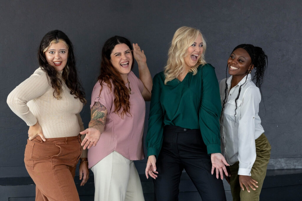 A group of four diverse women from a marketing agency standing together in a fun and energetic pose, showcasing camaraderie and reflection in their expressions.