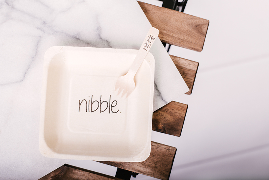 Eat Drink Host styled table setting featuring minimalist disposable plate and wooden utensil on marble surface.