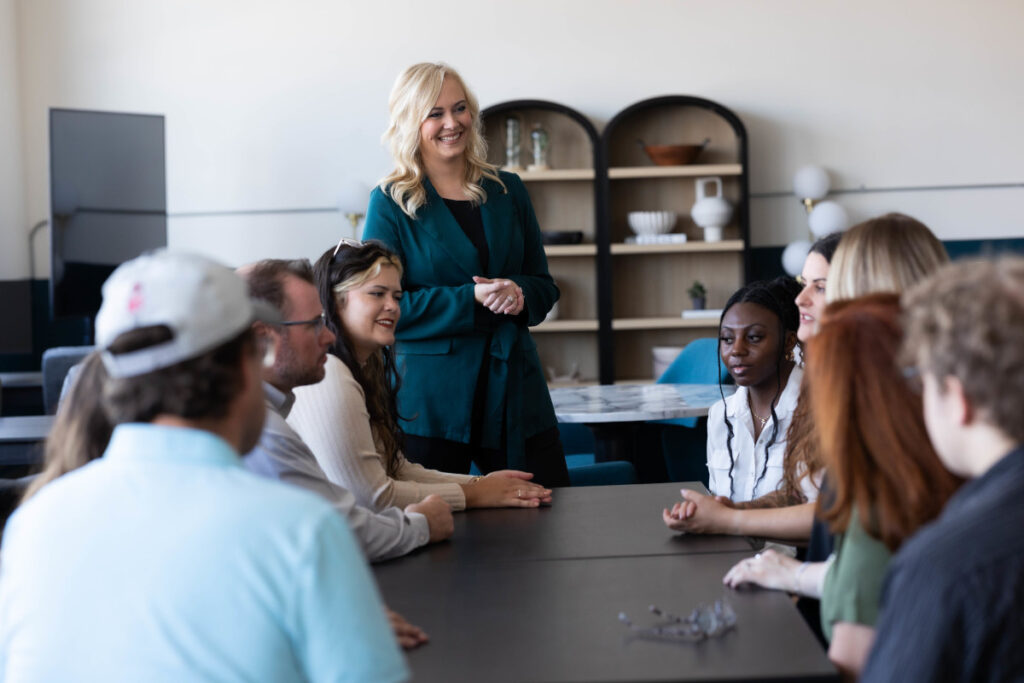 Team gathered around a table during a marketing education session led by a smiling instructor standing at the head of the group.
